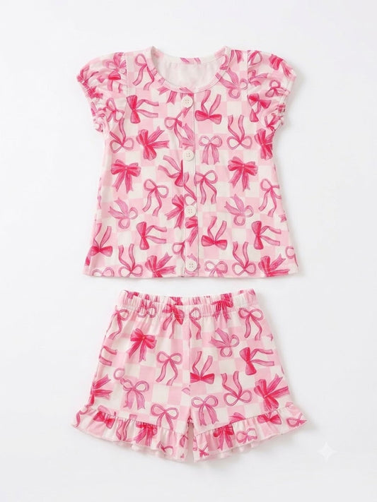 Girls 2-piece pink bow matching set featuring a short-sleeve button-front top with puff sleeves and an all-over pink bow print on a light pink background, paired with coordinating elastic waist shorts with ruffle hem