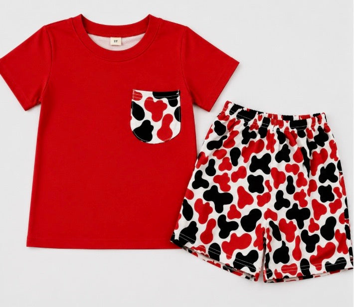 A children's two-piece clothing set featuring a solid red short-sleeve t-shirt with a patterned chest pocket, and matching shorts with a red, black, and white cow print.